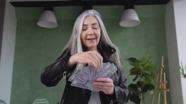 Close up of happy beautiful rich mature woman with gray hair which holding dollars bills like as handheld fan