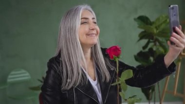Smiling satisfied mature woman with gray hair dressed in black jacket holding red rose in hand and making selfie
