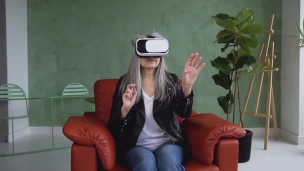 Portrait of charming modern focused 60-aged woman with gray hair which sitting in the armchair in special virtual 3d goggles — Stock video