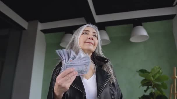 Крупним планом приваблива задоволена стильна старша жінка, яка махає доларами банкноти на фоні кімнати — стокове відео