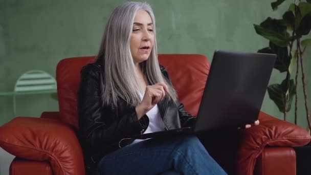 Mujer elegante sonriente de buen aspecto con el pelo gris sentado en una silla suave y tener video chat en el ordenador — Vídeos de Stock