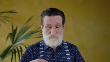 Front view of respected balanced bearded senior man which putting on his glasses and looking at camera on yellow background