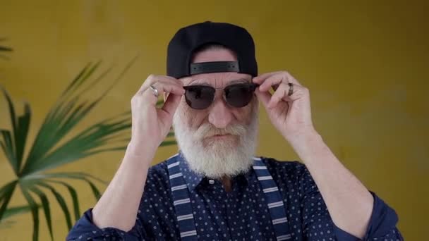 Front view of good-looking smiling satisfied senior bearded man in youthful cap taking off his glasses while looking at camera — 图库视频影像