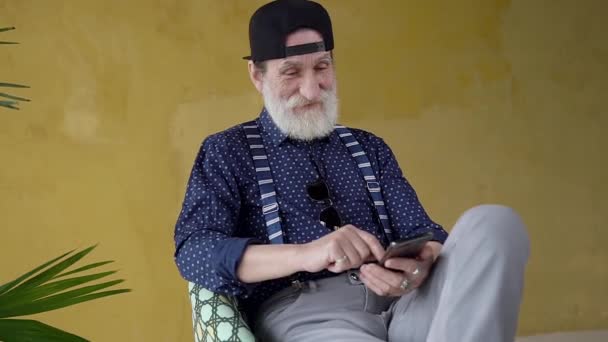 Rispettoso uomo anziano barbuto sorridente in abiti casual e in elegante berretto seduto sulla sedia e utilizzando il telefono vicino alla parete gialla — Video Stock