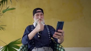 Front view of funny happy attractive bearded senior man which having video chat on phone ,sitting in chair on yellow background