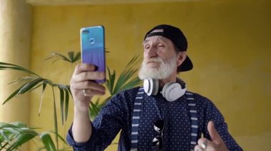 Attractive satisfied modern senior man with gray beard in trendy cap looking in phone camera to choose the best aspect to make photo