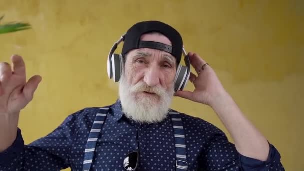 Красивий витягнутий стильний бородатий 70-річний чоловік в навушниках слухає красиву музику і дивиться на камеру — стокове відео