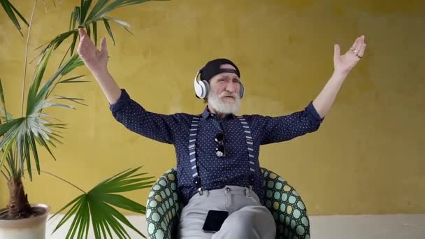 Front view of satisfied happy bearded retired man which sitting in chair near yellow wall with palm tree and enjoying the under songs in earphones — Stok video