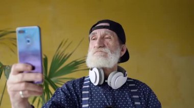 Concentrated attractive bearded mature man in stylish cap standing on yellow background and looking for the best look to make selfie