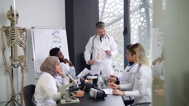 Retrato del médico cirujano sénior que tellinglecture para los colegas masculinos y femeninos concentrados multirraciales que sentaban la mesa de trabajo en la oficina del hospital — Vídeos de Stock