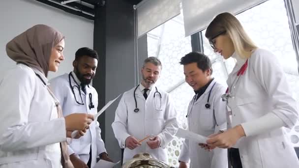 Portrait of professor and young medical interns of mixed race in medical clothing studying together human skeleton anatomy at lectures of surgery in light office in hospital — 图库视频影像