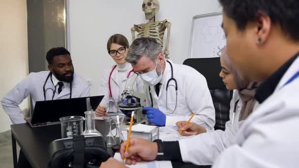 Group of young interracial scientists sitting at work table working with a microscope, test tubes and laptop in the laboratory — Stock video