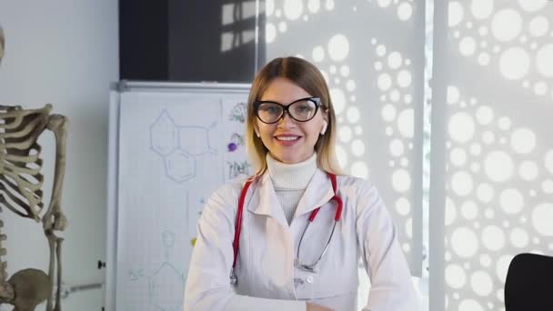Молодая женщина-врач в очках и белом халате со стетоскопом на шее улыбается в камеру в больничном кабинете. Врач, здравоохранение, любовь к медицине — стоковое видео