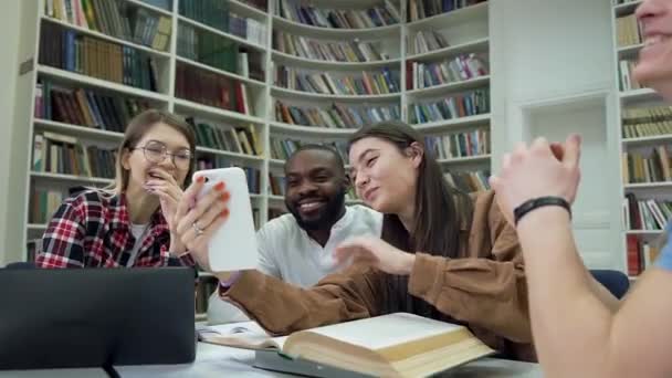 Atraente jovens multirraciais felizes sentados na biblioteca e sorrindo de vídeos engraçados no telefone meninas — Vídeo de Stock