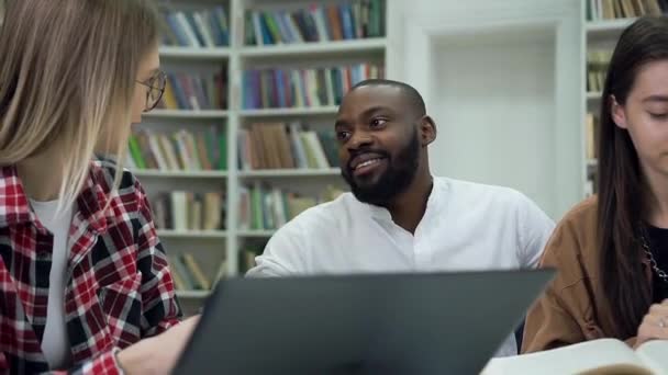 Beau sourire barbu afro-américain étudiant parlant avec sa compagne tandis qu'un autre leurs amis lecture livre dans la bibliothèque — Video