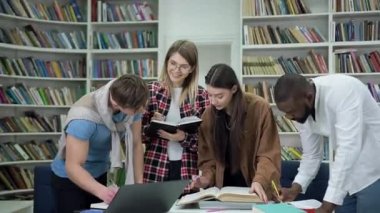 Attractive cheerful modern mixed race students preparing for exams in the university library and making needed notes into copybook