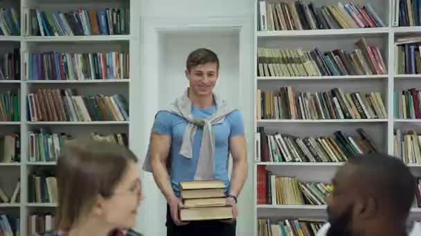 Front view of smiling handsome 25-aged guy which carrying a lot of books to his joyful attractive multiracial university friends waiting for him at the table — Stock video