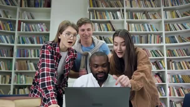 Atraente pessoas multiétnicas alegres bem sucedidas que satisfizeram da boa notícia na tela de computador portátil no contexto de biblioteca — Vídeo de Stock