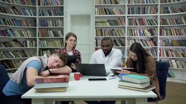 Vista frontal do cara bonito cansado que dorme na mesa e três outros estudantes multirraciais focados em boa aparência trabalhando no laptop na biblioteca — Vídeo de Stock