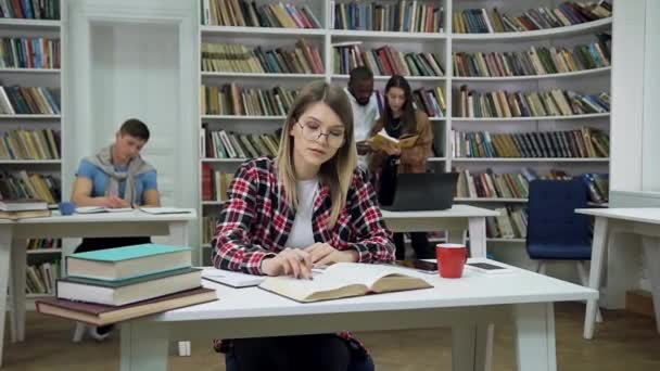 Front view of attractive focused blond young female student which sitting in the library, reading book and making notes — стоковое видео