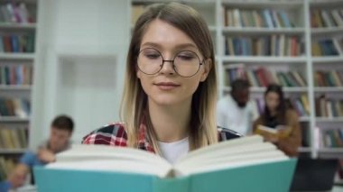 Pretty calm concentrated young woman in glasses reading book in the university library