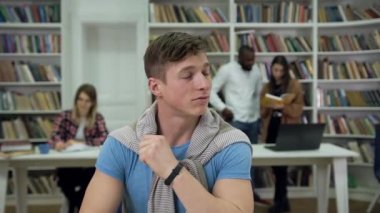 Thoughtful likable smart young man looking aside in the library and then writing notes into copybook on the another students background