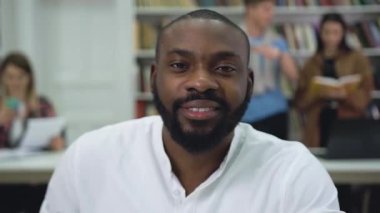 Good-looking high-spirited positive bearded young black-skinned man looking at camera in the library