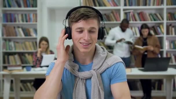 Joyful attractive modern young man in casual clothes sitting in the library and listening music in earphones — Stock video