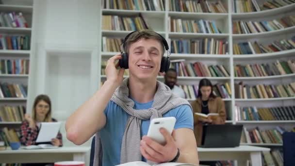 Повільний рух красивого задоволеного стильного хлопця в навушниках, який слухає музику, сидить в університетській бібліотеці — стокове відео