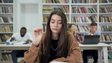 Front view of serious beautiful 25-aged modern woman with long hair which sitting in the library and reading book on the background of another diverse readers