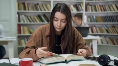 Charming focused modern young woman with long hair reading book in the university library and then looking aside with different thoughts
