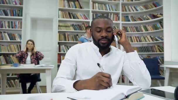 Rallentatore di simpatico ragazzo afroamericano barbuto soddisfatto che prende appunti in copybook durante la sua conversazione telefonica in biblioteca — Video Stock