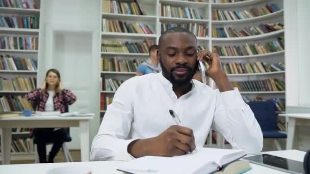 Cámara lenta de simpático chico afroamericano barbudo satisfecho que toma notas en copybook durante su conversación telefónica en la biblioteca — Vídeo de stock