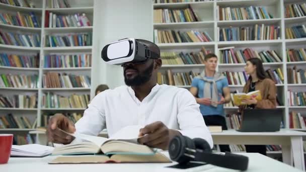 Piacevole concentrato barbuto giovane uomo dalla pelle scura seduto nella libreria con auricolare realtà virtuale e sfogliando un libro — Video Stock