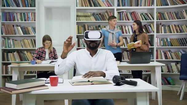 Vue de face d'attrayant jeune homme barbu concentré à la peau foncée dans des lunettes spéciales 3D qui bouge les mains sur un écran imaginaire, assis dans la bibliothèque — Video