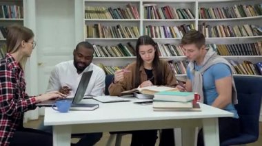 Portrait of likable creative diverse young girls and guys which sitting together at the table in the library and doing home task