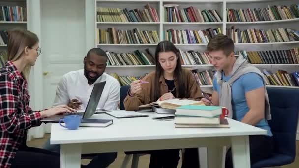 Portrait of likable creative diverse young girls and guys which sitting together at the table in the library and doing home task — ストック動画