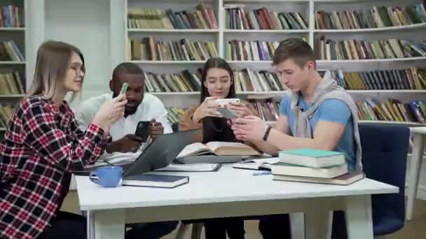 Attraente allegro giovani amici multirazziali che giocano sui loro telefoni durante la pausa tra la preparazione per gli esami in biblioteca — Video Stock