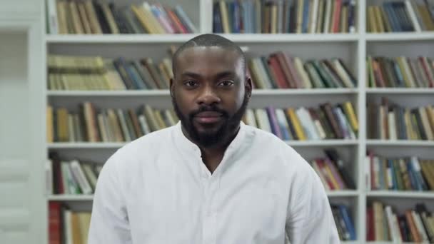 Atractivo sonriente joven barbudo afroamericano en camisa blanca de pie cerca de las estanterías de la biblioteca y mirando a la cámara — Vídeo de stock