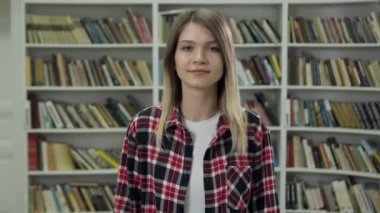 Cute smiling modern young woman in casual clothes standing near shelves with different books in the library and posing on camera