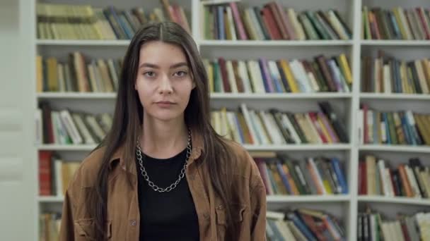 Vista frontal da menina 25-aged concentrada bastante confiante com cabelo marrom que olha para a câmera perto de estantes na biblioteca — Vídeo de Stock