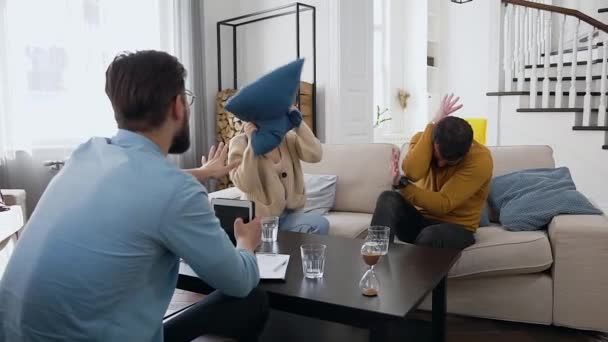 Verängstigter junger bärtiger Mann wehrt sich dagegen, ihn mit einem Kissen zu schlagen, das seine wütende Freundin während eines Besuchs beim Psychologen hält — Stockvideo
