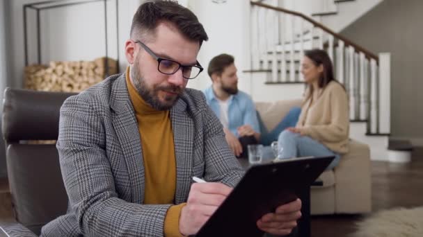 Přední pohled na seriózní profesionální vousatý psycholog, který si dělá poznámky na pozadí usmívající se mladý manželský pár, který mluví — Stock video