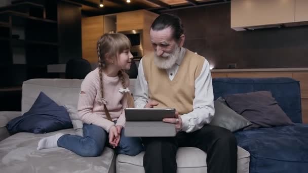 Смолится счастливая симпатичная 12-летняя девочка, сидящая на диване со своим уважаемым бородатым старшим дедом и что-то просматривающая на i-pad — стоковое видео