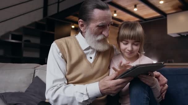 Aantrekkelijke gelukkige oudere bebaarde man zit op de bank samen met zijn vrolijke mooie tiener kleindochter en met behulp van i-pad — Stockvideo