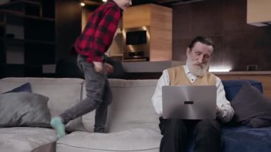 Gürültülü, gülümseyen 10 yaşında bir çocuğun ön görüntüsü. Ciddi, olgun, sakallı büyükbabasının yanına atlıyor ve bilgisayarda çalışırken ona müdahale ediyor.