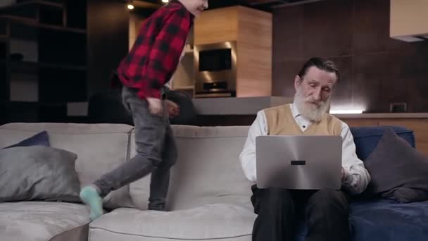 Front view of noisy smiling 10-aged boy which jumping near his serious mature bearded grandfather and interfering him while he working on computer — Stock Video