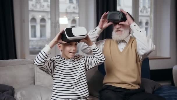 Zeitlupe eines verspielt freundlich lächelnden bärtigen Seniors zusammen mit seinem Teenager-Enkel beim Videospiel mit spezieller 3D-Brille zu Hause — Stockvideo