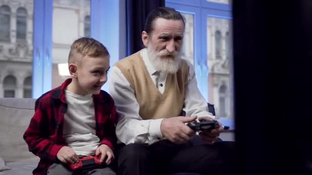 Movimento lento de bom aspecto feliz sorridente barbudo homem sênior, juntamente com seu neto alegre de 10 anos jogando videogame usando joysticks — Vídeo de Stock