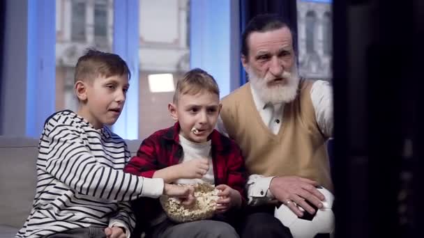 Frontansicht zweier ernsthafter Teenie-Jungs, die Popcorn essen und mit ihrem älteren bärtigen Opa das Sportspiel im Fernsehen diskutieren — Stockvideo